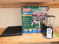 Coleman Party Grill box, lantern, and cast iron grate on shelf