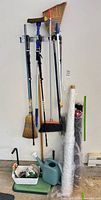 Wall-mounted assortment of brooms, poles, rolls of wrap, watering can, caddy with brushes