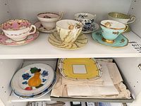 Shelf view showing five teacup & saucer sets, decorative plates and linens