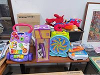 Overview of lot on table showing walker, microphone box, spinning toy, crib soother, water blaster and bin of plush