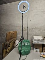Ring light on tripod illuminated, power cable visible, green screen bag stored below