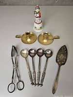 Entire lot laid out: spoons, tongs, pie server, brass candle holders, ceramic bell