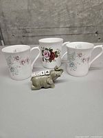 Front view of two Royal Devon floral teacups, one Queen's Centenary rose teacup, and grey elephant perpetual calendar