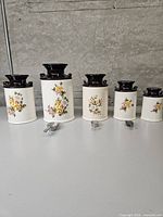 Five ceramic canisters with brown lids arranged by size, three metal scoops placed in front