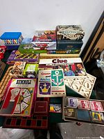 Overview of stacked boxed games, visible titles include Rummoli, Probe, Scene It?, Crown & Anchor, Clue board, decks of cards, and loose racks
