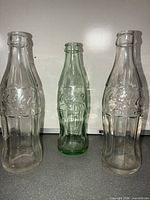 Front view of three Coca-Cola contour bottles showing clear and green glass and embossed logos