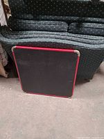 Overall view of square black card table top with red trim leaning against sofa, shows surface and edges