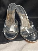 Front angled view of both clear mule heels showing transparent uppers and branding on insole