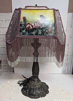 Full view of lamp showing ornate metal base and beaded fringe shade with painted rose panel