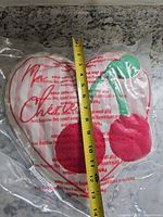 Front view of heart-shaped pillow in plastic wrap with cherries and script, tape measure showing height (~10 in)