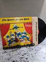 Front cover and partially pulled black vinyl disc for The Smurfs All Star Show