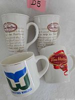 Four white ceramic mugs with Tim Hortons and Hartford Whalers logos arranged against wall