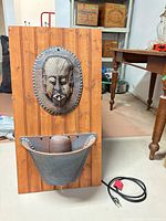 Full view of wall fountain showing wood backboard, mask spout, basin and power cord