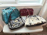 Four handbags on table: teal shoulder bag, black metallic satchel, burgundy handbag, cream floral tote