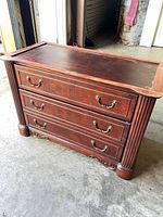 Front view showing three drawers, brass-tone pulls, fluted sides, carved base