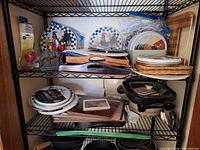 Shelf showing rooster burner covers, wooden boards, electric skillet, woks, placemats