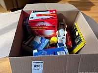Open cardboard box showing multiple boxed light bulbs including Pricemark 40 W 12-pack, loose yellow bulb, various other boxes