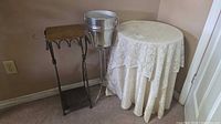 Full view of three items: round accent table with cloth, metal champagne bucket on stand, and metal plant stand