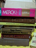 Stack of six books showing spines including The Merck Manual, Wild Plants of the Canadian Prairies, Warrior’s Woman, Clear and Present Danger, The Warlord