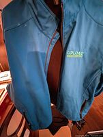 Front of blue Upload softshell jacket hanging, showing zipper and logo