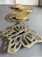 Four metal trivets: three round apple-shaped brass pieces and one butterfly trivet