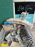 Overall lot view showing cutting mat, tote with buttons, trims, patterns, satin hangers, fringe tablecloth and boxed placemat set