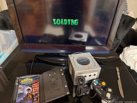 Console, cables, controller, empty game case displayed under powered TV showing loading screen