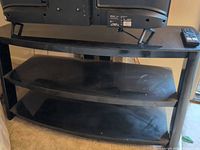 Front view of three-tier black glass TV stand with TV on top and remote control on upper shelf
