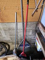 Three long-handled garden tools leaning against wall: straw broom, Kobalt leaf rake, red-handled push broom