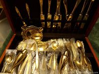 Photo showing an array of gold-tone flatware including knives, forks, spoons and serving pieces arranged in a storage drawer. Most pieces individually wrapped in plastic.