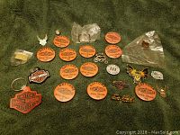 Photo showing a flat lay of multiple Harley Davidson and motorcycle-related pins and keychains on a green textured surface for display.