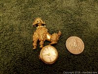 Front view of gold-tone poodle brooch with small attached white-faced watch, shown next to a quarter for scale.