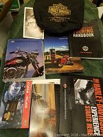 Items displayed on a green cloth including various Harley-Davidson books and pamphlets along with a black motorcycle cover embroidered with Harley-Davidson logo.