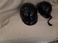 Two black motorcycle helmets displayed on a beige fabric surface, showing their outer shells with visible scratches.