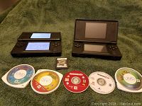 Two purple Nintendo DS Lite consoles opened and powered on viewed from top, with five PSP game discs and one Pokemon Black DS cartridge arranged in front.