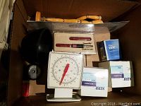 Box interior showing wooden mannequin lying on top of other items along with a white mechanical scale and several boxes of medical supplies.