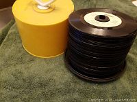 Stack of approximately 50 45 RPM vinyl records on a spindle with a yellow plastic storage case beside them.