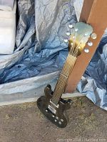 Black guitar-shaped decorative item resting against a post on dirt ground with a tarp in the background. Visible tuning pegs, strings, and control knobs present, dust visible on the surface.