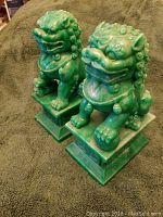 Side angle view of two green Foo Dog statues with curled manes and pedestal bases on a dark cloth.