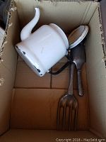 Photo of a cardboard box containing a white enamel teapot, a metal spatula, and a large metal fork, showing signs of wear and age.