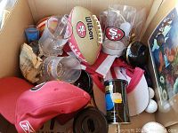 Photo shows a box of assorted sports memorabilia, prominently featuring a Wilson branded San Francisco 49ers football, drinking glasses with gold rims, a red 49ers cap, golf balls, a can of beer, and other related items.