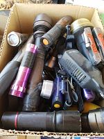 Photo showing multiple flashlights tightly packed in a cardboard box. Flashlights vary in color and size including purple, black, brown, and blue. Lantern partially visible.