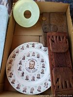 A cardboard box contains two round ceramic plates featuring portraits of US Presidents arranged in a circular pattern and a light green ceramic dish with a hole in its center, along with a brown leather pouch and a carved wooden elephant bookend partially visible.