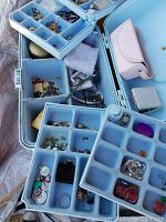 Overview of multiple blue compartmentalized boxes containing assorted small beads, buttons, and jewelry findings, plus a small pink jewelry case and small gray box