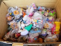 Cardboard box filled with numerous McDonald's Happy Meal toys in original plastic bags with various colors and shapes visible, showing condition and quantity.