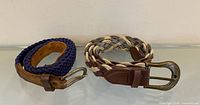 Two belts coiled, showing woven straps, leather ends and brass buckles