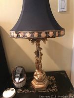 Side view of the lamp showing the male figurine base in gold finish with hexagonal stepped base, remote controls nearby for scale.