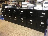 Photo showing six black two-drawer file cabinets side by side with documents and storage boxes on top.