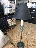 Single tall candlestick lamp with black paper shade, showing base and partial column with patina/wear.