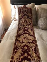 Full view of the velvet table runner laid out on a bed, showing burgundy fabric with a gold floral design and tassel ends.